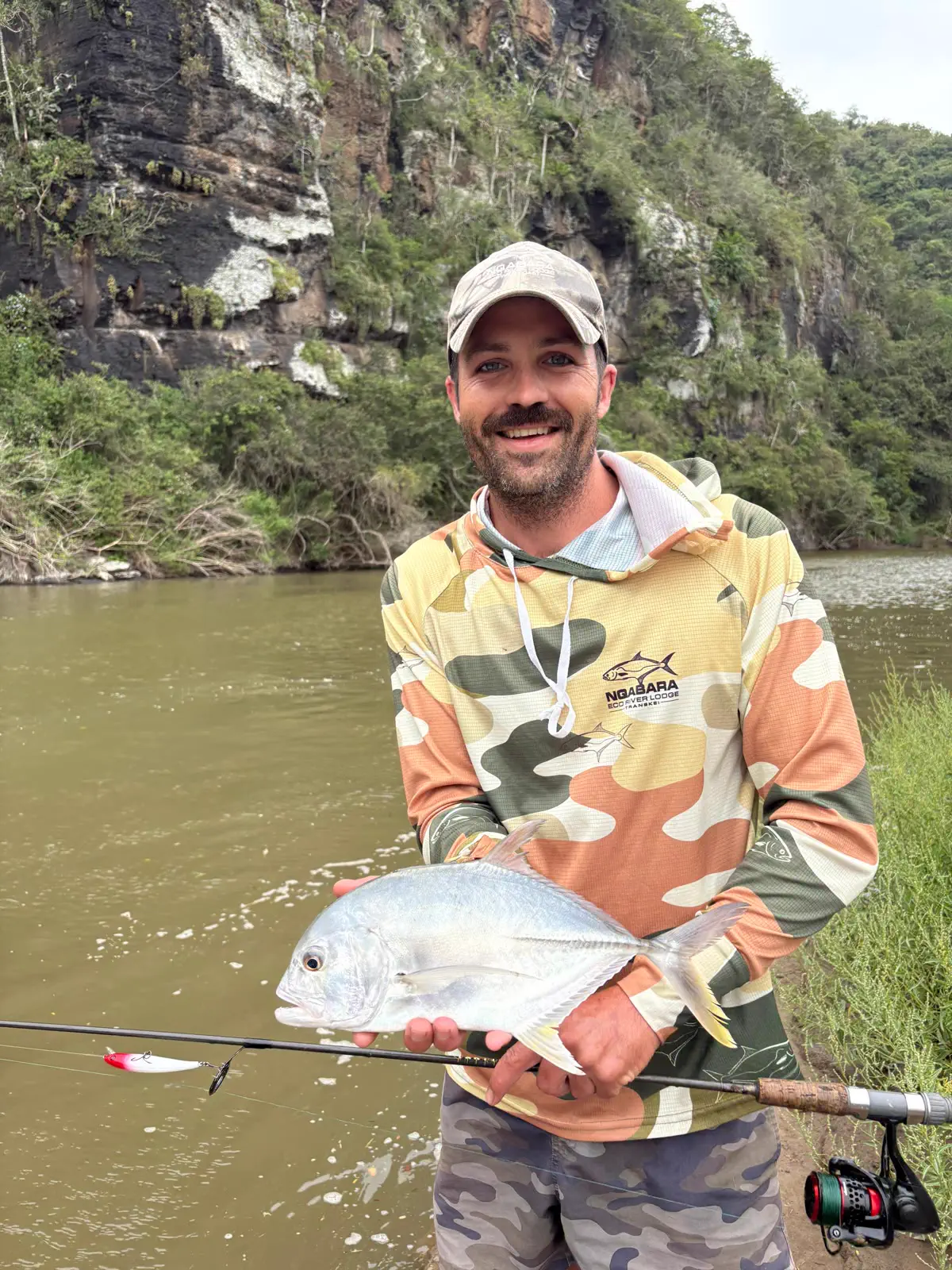 An angler below the river cliffs holding a silver fish and a fishing rod